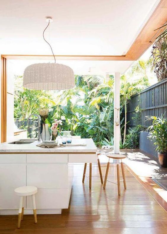 sleek white kitchen island, a white wicker lampshade, wooden stools and a tropical garden, to which the kitchen is opened
