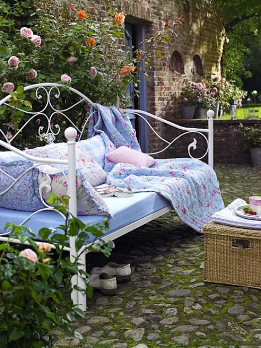 A vintage inspired white forged daybed with many pillows and a floral blanket is timeless classics for an elegant garden