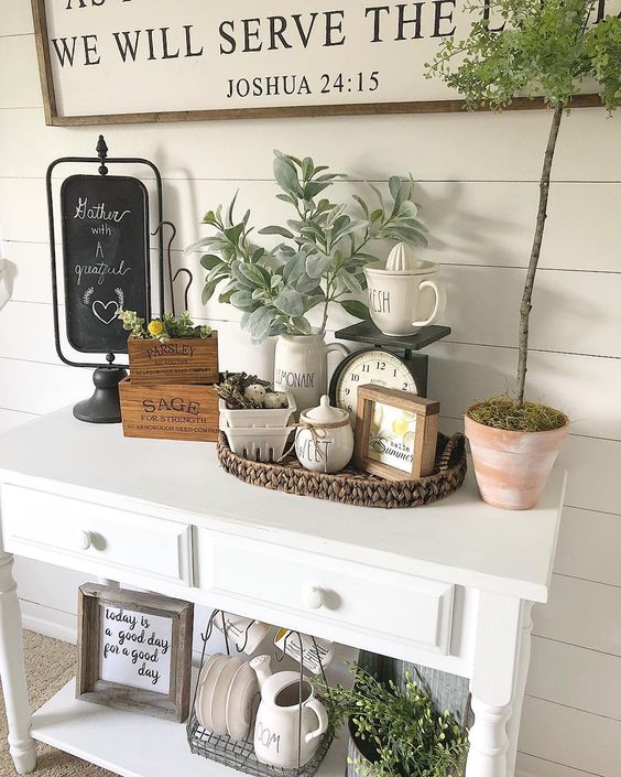 a cozy summer farmhouse entryway with a sign, pale greenery, a basket, some pots and porcelain