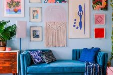 a bold and chic living room with pale blue walls, bold blue seating furniture, a stained dresser, a bright gallery wall and a retro chandelier