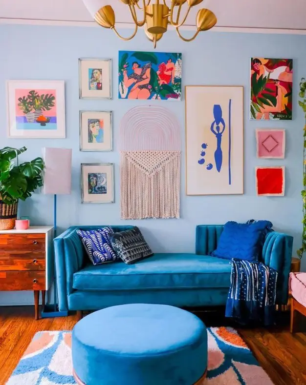 a bold and chic living room with pale blue walls, bold blue seating furniture, a stained dresser, a bright gallery wall and a retro chandelier