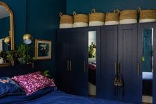 a bold eclectic bedroom with navy walls, navy wardrobes, baskets, a bed with electric blue bedding, a mirror and gold lamps