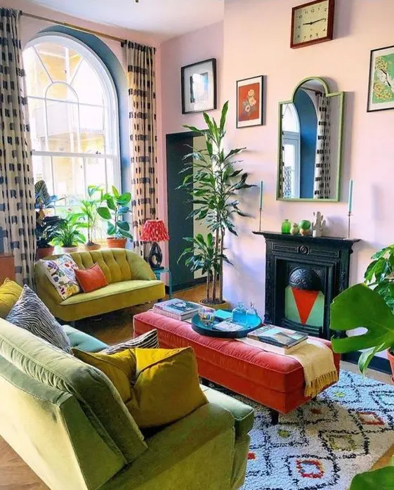 A bright maximalist living room with pink walls, mustard and neon green furniture, a vintage fireplace, a red ottoman and some statement plants.
