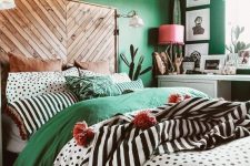 a catchy eclectic bedroom with emerald walls, a bed with a wooden headboard, colorful and printed bedding, a beaded chandelier, a pink lamp and some cacti