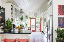 a catchy eclectic kitchen with white cabinets, bold red stools and doors, pendant lamps and potted greenery