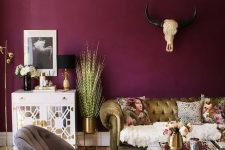a catchy eclectic living room with a purple accent wall, a gold sofa and a grey chair, a coffee table, a mirror cabinet and a bold gold chandelier