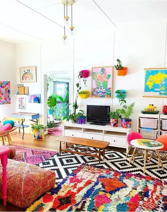 A colorful maximalist living room with bold layered rugs, pink printed cushions on the floor, some colorful chairs and artworks.