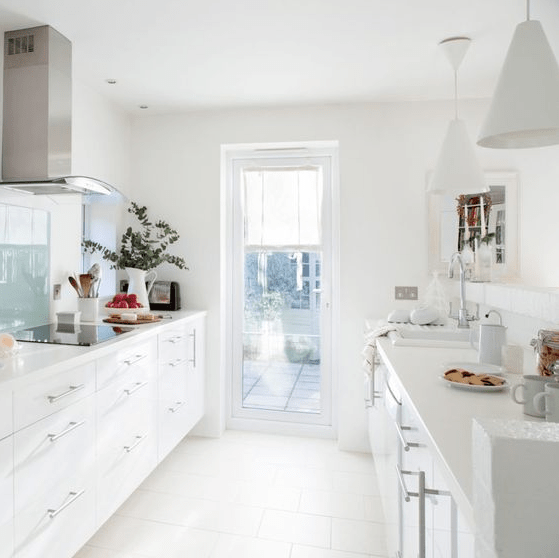 a contemporary white galley kitchen with white cabinets, metal and glass touches and an access to the terrace with much light