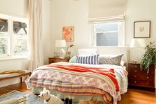 a cozy eclectic bedroom with a bed and bright bedding, stained nightstands, artwork, greenery and a chair