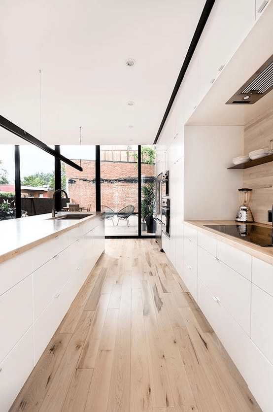 A cozy minimalist galley kitchen in white, with light colored wooden countertops and a floor plus a backsplash, with glazed walls for more light