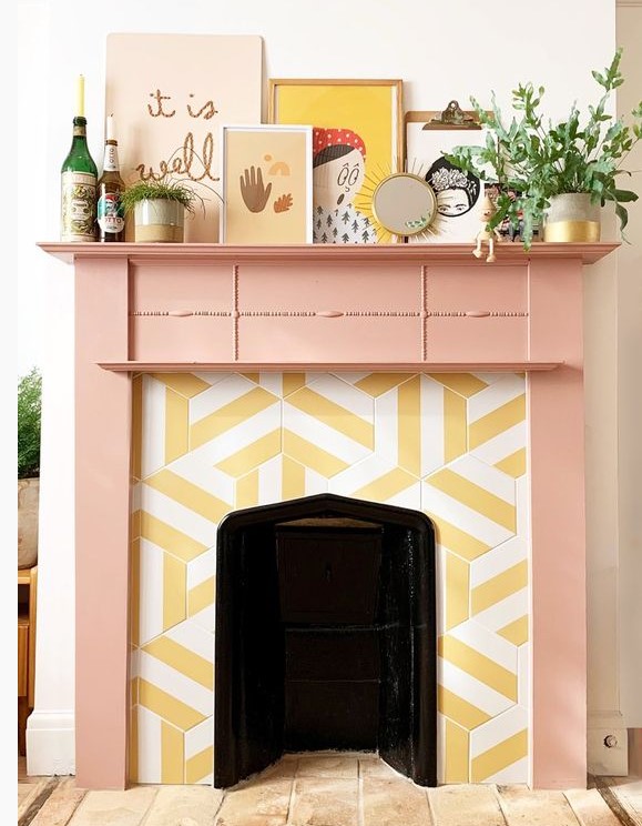 a faux fireplace with yellow and white graphic tiles around it and a peachy pink frame and mantel with a qurky gallery wall