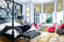 a gorgeous maximalist bedroom with lilac walls, a black bed and an ottoman, red chairs and a grey windowsill bench, a crystal chandelier