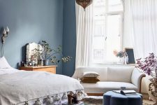 a lovely eclectic bedroom with navy walls, a bed with neutral bedding, a dark green bench, a white loveseat and a Moroccan lamp