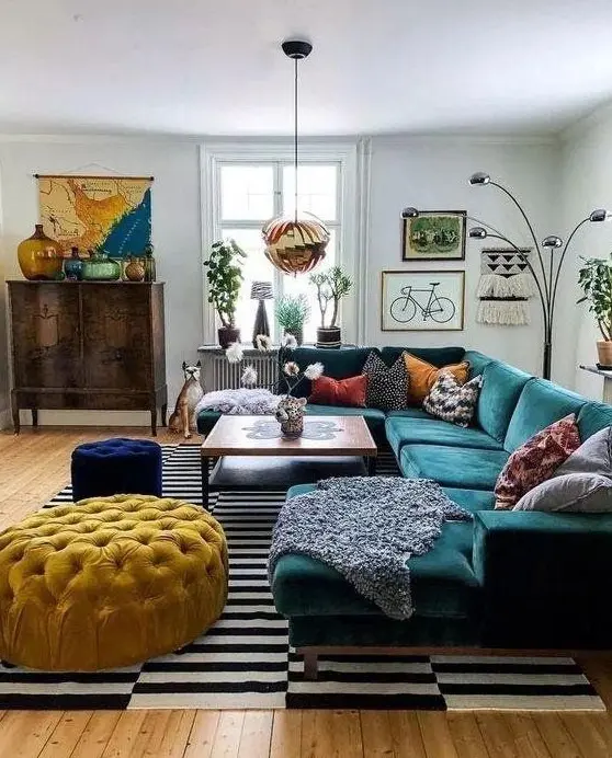 a lovely eclectic living room with a green sectional, a coffee table, a navy stool and a yellow ottoman, a stained credenza and artwork
