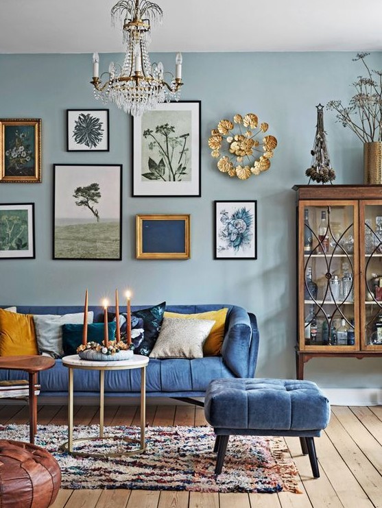 a lovely eclectic living room with light pink walls, a blue sofa and a pouf, a stained table, a beautiful gallery wall and a crystal chandelier