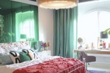 a maximalist bedroom with simple furniture, green mirrors and curtains, a colorful rug and bold bedding plus a fluffy chandelier