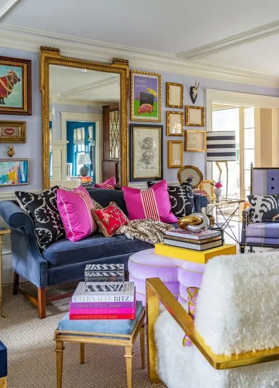 A maximalist living room with lilac walls, a navy sofa, a purple heart ottoman, colorful pillows and books and a gallery wall with a large mirror.