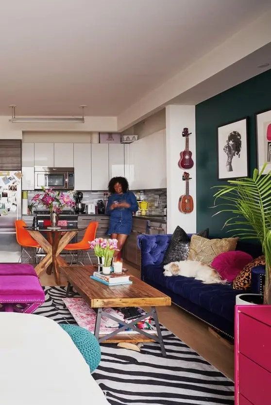 A maximalist open layout with a dark green accent wall, a navy sofa, colorful printed pillows, hot pink stools and a printed rug.