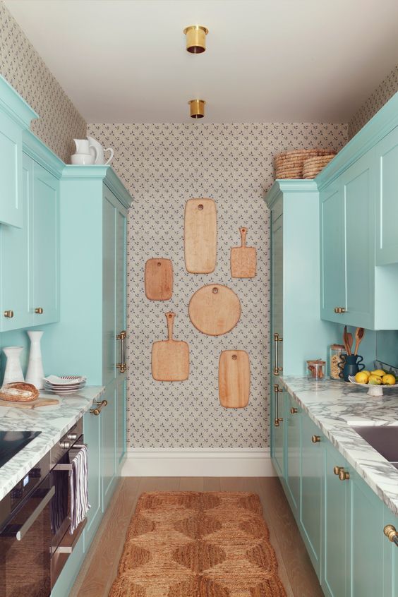 a mint blue galley kitchen with marble countertops, a gallery wall of cutting boards and a jute rug is cozy and cute