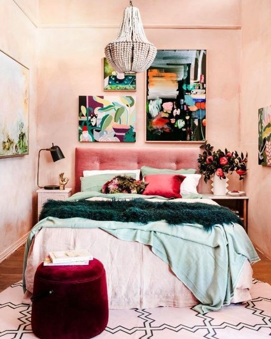 a modern eclectic bedroom with blush textural walls, a pink upholstered bed, colorful bedding, a burgundy pouf, a beaded chandelier and a colorful gallery wall