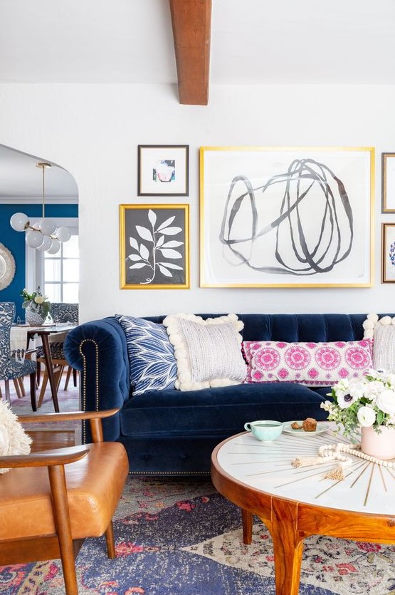 A modern eclectic living room with a navy sofa, amber chairs, a round coffee table, colorful pillows, a free form gallery wall.