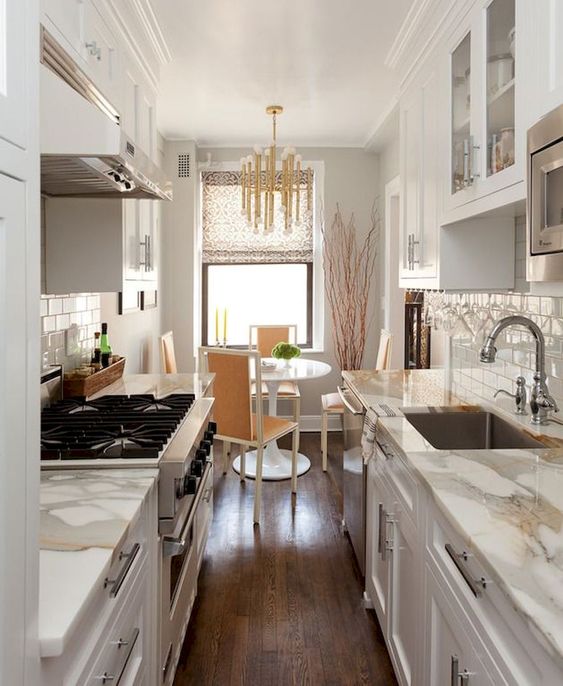 a small and welcoming galley kitchen with white cabinets and white stone countertops, a cozy dining space wth a round table and a glam chandelier