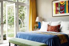an eclectic and colorful bedroom with tan walls, a bed with colorful bedding, a green bench, a colorful boho rug and yellow curtains