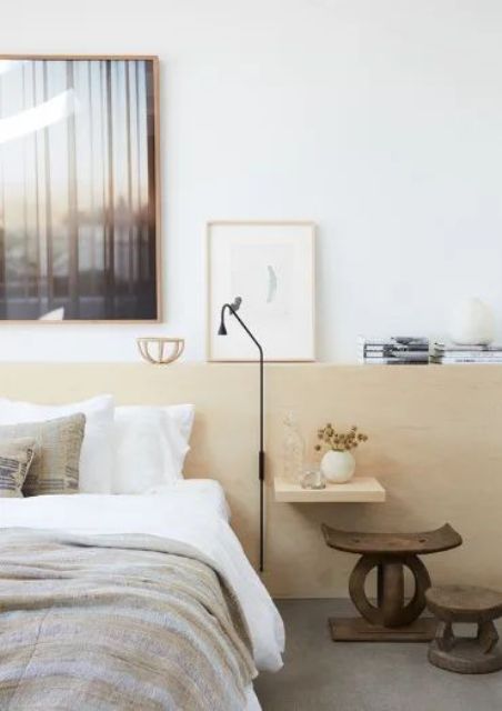 An eclectic bedroom with a bed and neutral bedding, a stained headboard panel for storage, dark stained stools and a sconce