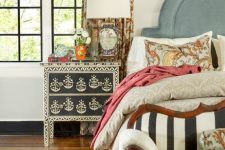 an eclectic bedroom with a blue upholstered bed, colorful bedding, a stenciled nightstand, a striped loveseat, printed curtrains