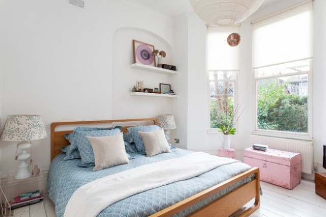 An eclectic bedroom with a modern wooden bed, a pink chest and stool, vintage inspired lamps and modern built in shelves