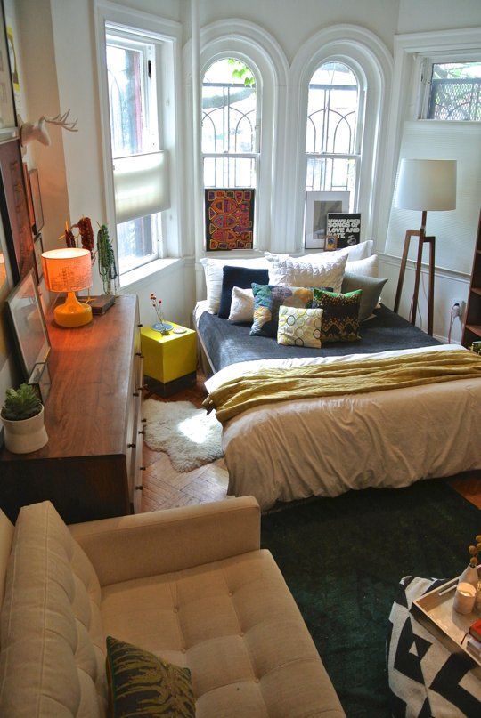 an eclectic bedroom with an arched window, a bed with bright pillows, a neutral chair, a stained dresser and a floor lamp