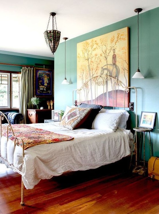 An eclectic bedroom with green walls, a forged bed with colorful and printed bedding, forged stools, a dark stained desk, a chic chandelier and an oversized artwork
