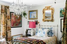 an eclectic guest bedroom with a metal bed and bright textiles, a pouf, a rattan chair, a gallery wall and a crystal chandelier
