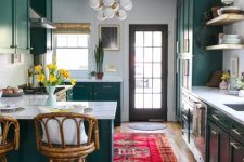 an eclectic kitchen done with emerald cabinets, white marble countertops, tall rattan stools, modern chandeliers and a bold boho rug