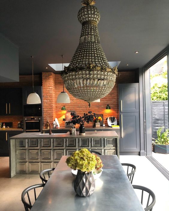 an eclectic kitchen with dark shaker style cabinets, a shiny kitchen island, a red brick wall, a dining zone with a metal table, black chairs and a chandelier