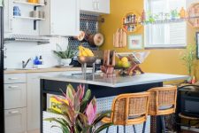 an eclectic kitchen with mustard walls, white cabinets and a black kitchen island, rattan stools, a crate and potted plants
