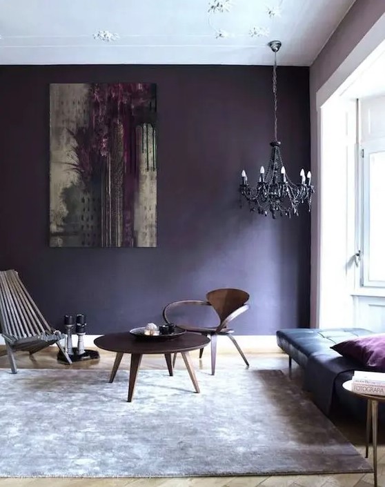 With a deep violet accent wall, this eclectic living room boasts a striking black leather daybed. A set of wooden chairs and a simple table under a bold black chandelier complete the look, making the space inviting and stylish.