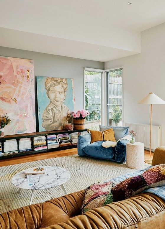An eclectic living room with a blue and mustard chair, a beige sofa, colorful pillows, a bookshelf, a marble table and artwork.
