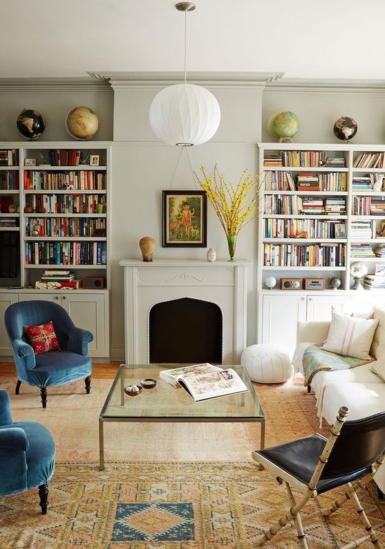 An eclectic living room with built-in bookshelves, a fireplace, a creamy sofa, blue chairs, a black one, a glass coffee table and printed rugs.