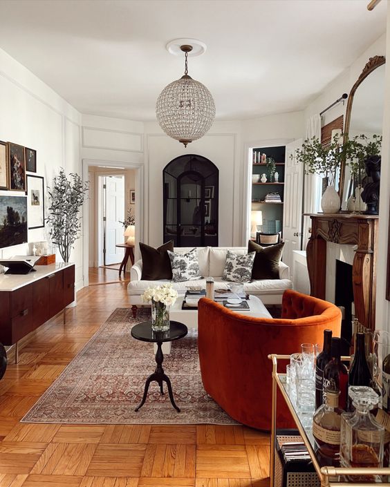 An elegant eclectic living room with a stained credenza, a rust chair, a white sofa, some nightstands, a crystal chandelier.