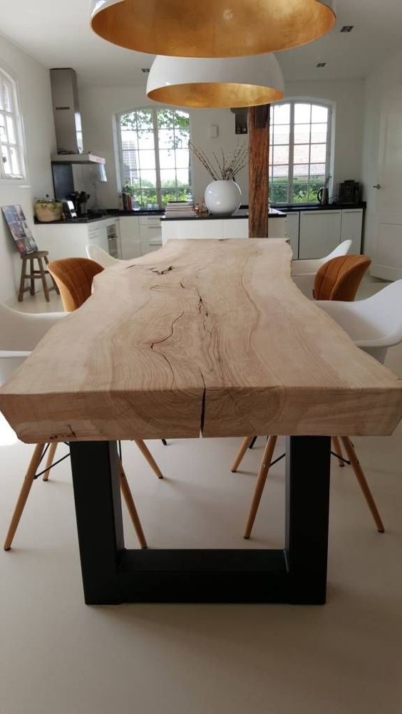 A light stained dining table of a single slab with a live edge and mismatching rust and white chairs