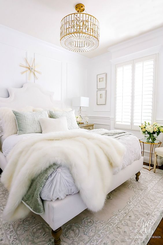 a neutral glam bedroom with white walls, a statement crystal chandelier and chic bedding plus faux fur