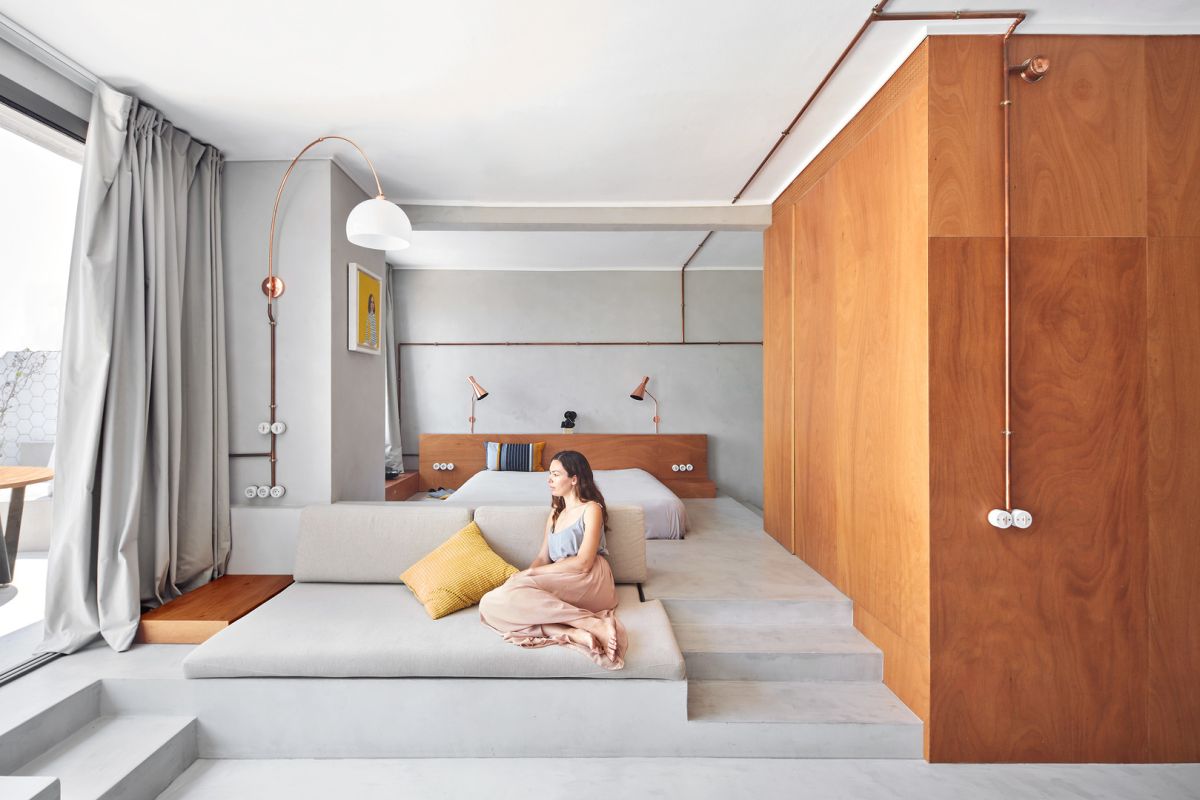 The bedroom features a bed on a platform, lamps and a large window and semalessly flows into a living room that flows into a terrace
