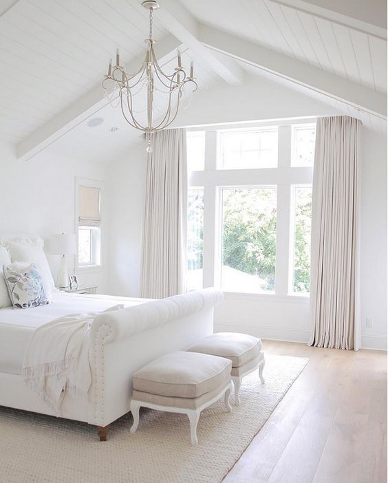 a very fresh white bedroom with creamy curtains and stools, an elegant chandelier and a white upholstered bed