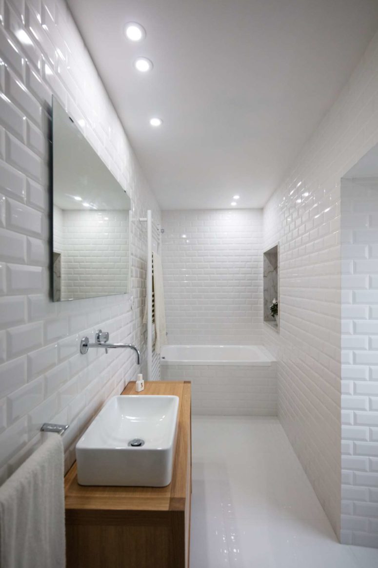 The bathroom is fully done in white, with chic tiles, a wooden vanity and a built-in bathtub