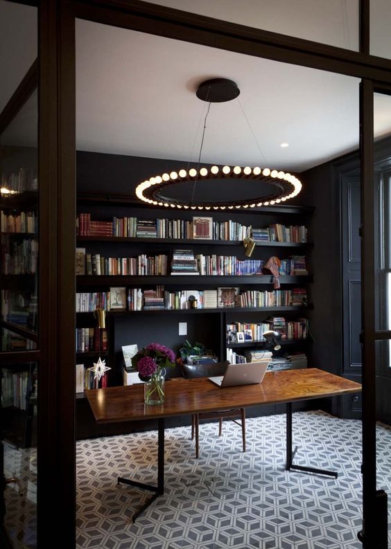 a black home office with bookshelves, a catchy modern chandelier, a wooden desk and a printed carpet