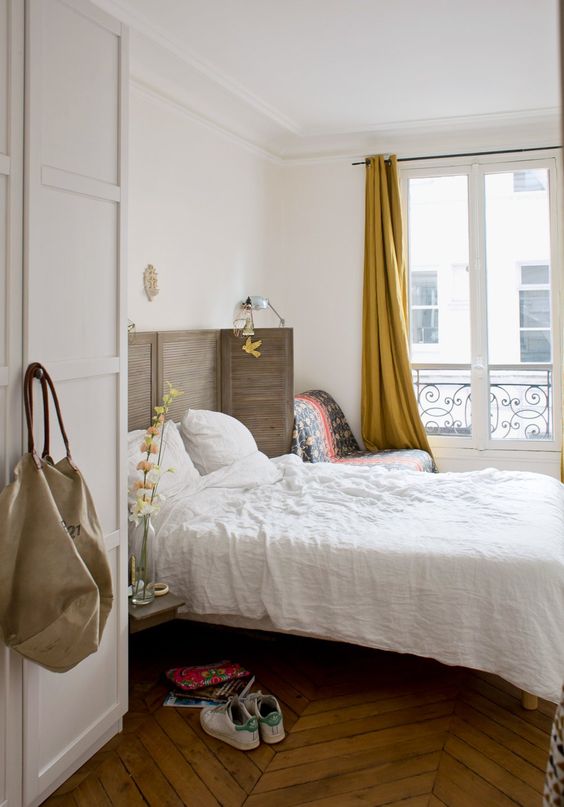 A Paris inspired chic bedroom with a shutter screen as a headboard, a herringbone floor, mustard curtains and a comfy chair