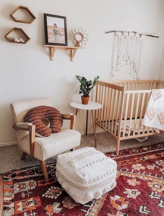 A bright boho nursery with a red boho rug, a white Moroccan pouf, macrame, mid century shelves and a chic chair