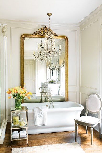 a chic Parisian bathroom with a statement mirror in a gilded frame, a crystal chandelier, a chic tub and vintage furniture