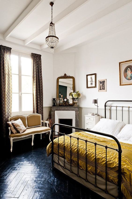 a chic Parisian bedroom with printed curtains, a crystal chandelier, a retro metal bed, a chic mustard loveseat and a marble clad fireplace